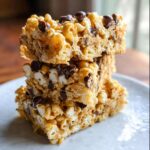 Three stacked squares of gooey Peanut Butter Marshmallow Popcorn bars topped with chocolate chips on a white plate.