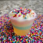 Close-up of a Sugar Cookie Jello Shot in a clear cup, topped with whipped cream and colorful sprinkles, surrounded by more sprinkles.