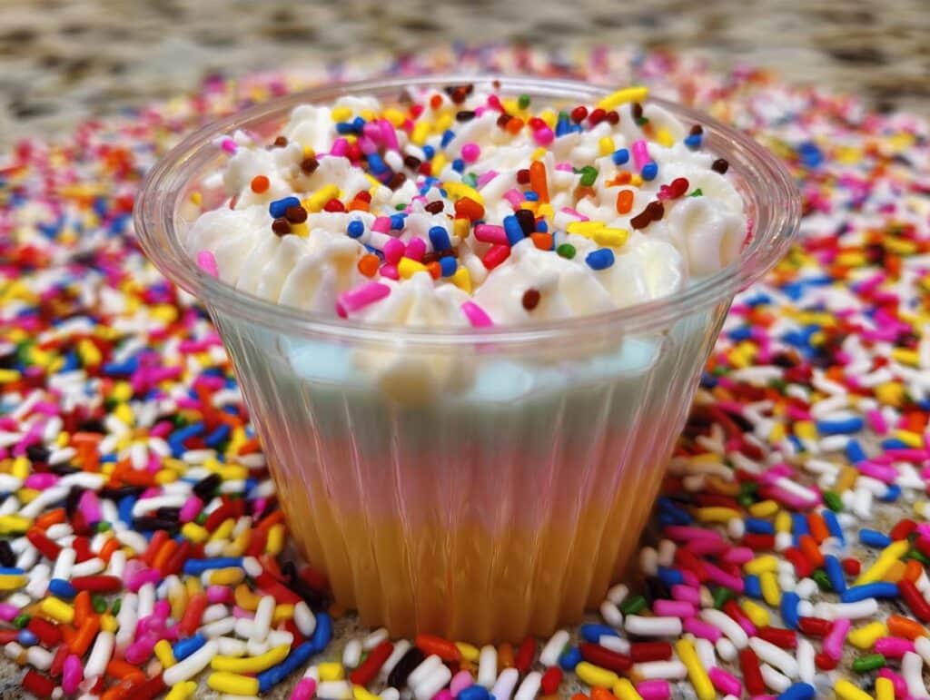 A close-up of a Sugar Cookie Jello Shot topped with whipped cream and colorful sprinkles, surrounded by a bed of sprinkles.