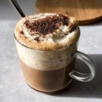 A close-up of a Tiramisu Latte in a glass mug, topped with whipped cream and cocoa powder.