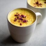 Close-up of a creamy Turmeric Golden Milk Latte in a white mug, garnished with dried rose petals and a sprinkle of cinnamon.