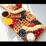 A vibrant Waffle Breakfast Board featuring waffles, fresh berries, sliced bananas, orange slices, nuts, and whipped cream.