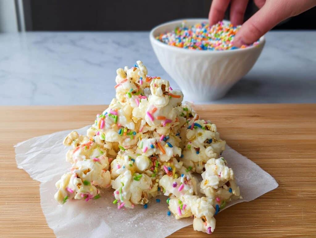 A pile of White Chocolate Funfetti Popcorn covered in colorful sprinkles on parchment paper.