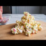 A pile of White Chocolate Funfetti Popcorn coated in white chocolate and colorful sprinkles on a wooden board.