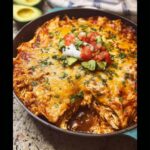 A close-up of cheesy 20-Minute Skillet Chicken Enchiladas topped with sour cream, pico de gallo, and cilantro in a cast iron pan.