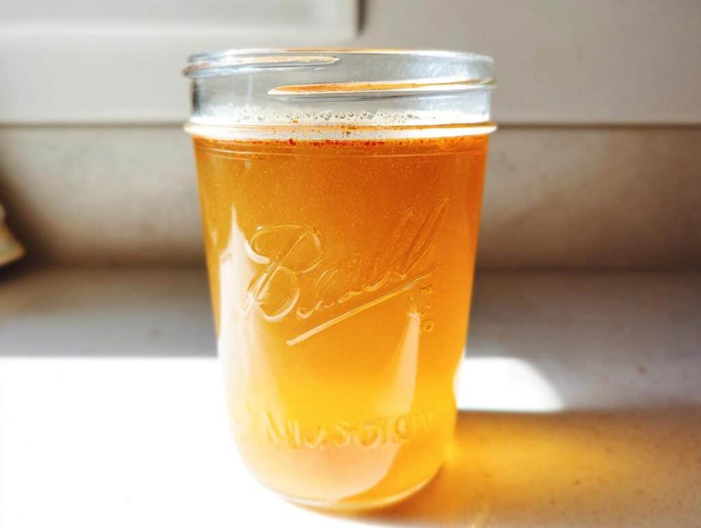 A glass Ball mason jar filled with an amber liquid, representing an Apple Cider Vinegar Detox Drink, sitting on a white surface.