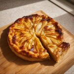 A golden brown Apple Free-Form Tart with thinly sliced apples arranged in a circular pattern, sitting on a wooden board with one slice removed.