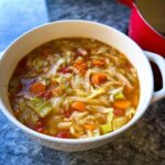 A close-up of a white bowl filled with hearty Cabbage Detox Soup featuring shredded cabbage, carrots, and tomatoes.