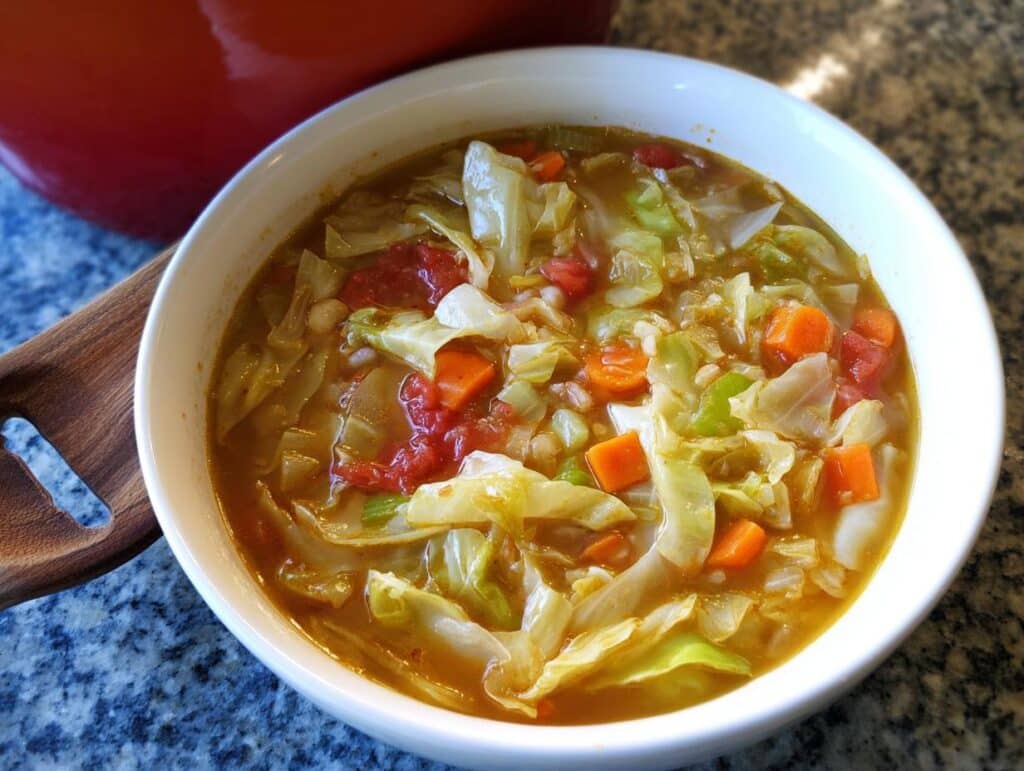 A close-up of a white bowl filled with hearty Cabbage Detox Soup, featuring large pieces of cabbage, diced carrots, and tomatoes.