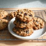 A stack of four chewy Oatmeal Raisin Cookies sits on a small white plate, with more cookies cooling on a wire rack.