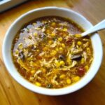 A close-up overhead shot of a white bowl filled with rich, dark Chicken Corn Hot & Sour Soup, featuring shredded chicken and yellow corn kernels.