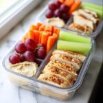 Close-up of a healthy Chicken Hummus Lunch Box featuring sliced seasoned chicken, carrots, celery, grapes, and hummus.