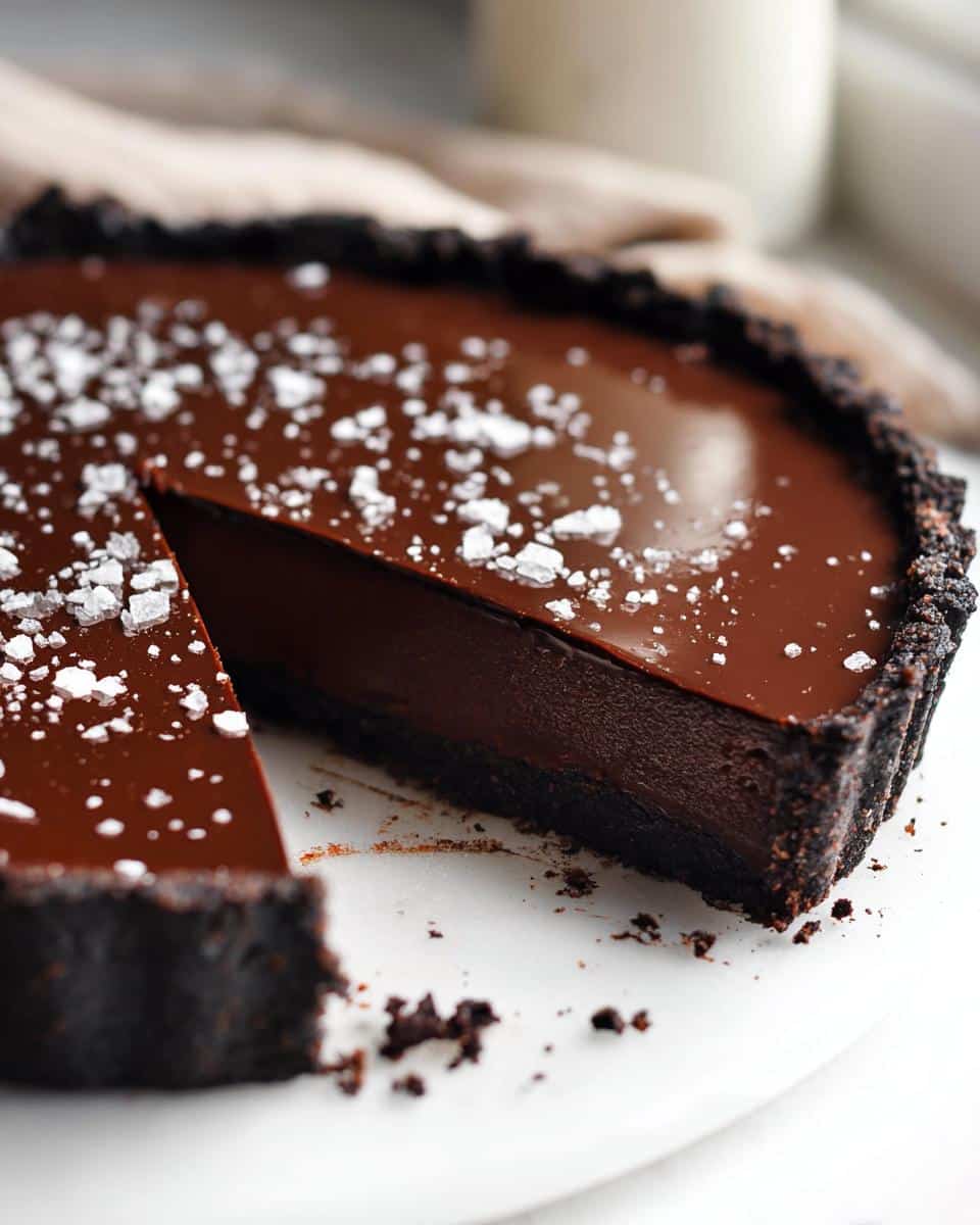 A decadent slice removed from a Chocolate Ganache Tart with a dark crust and flaky sea salt sprinkled on top.