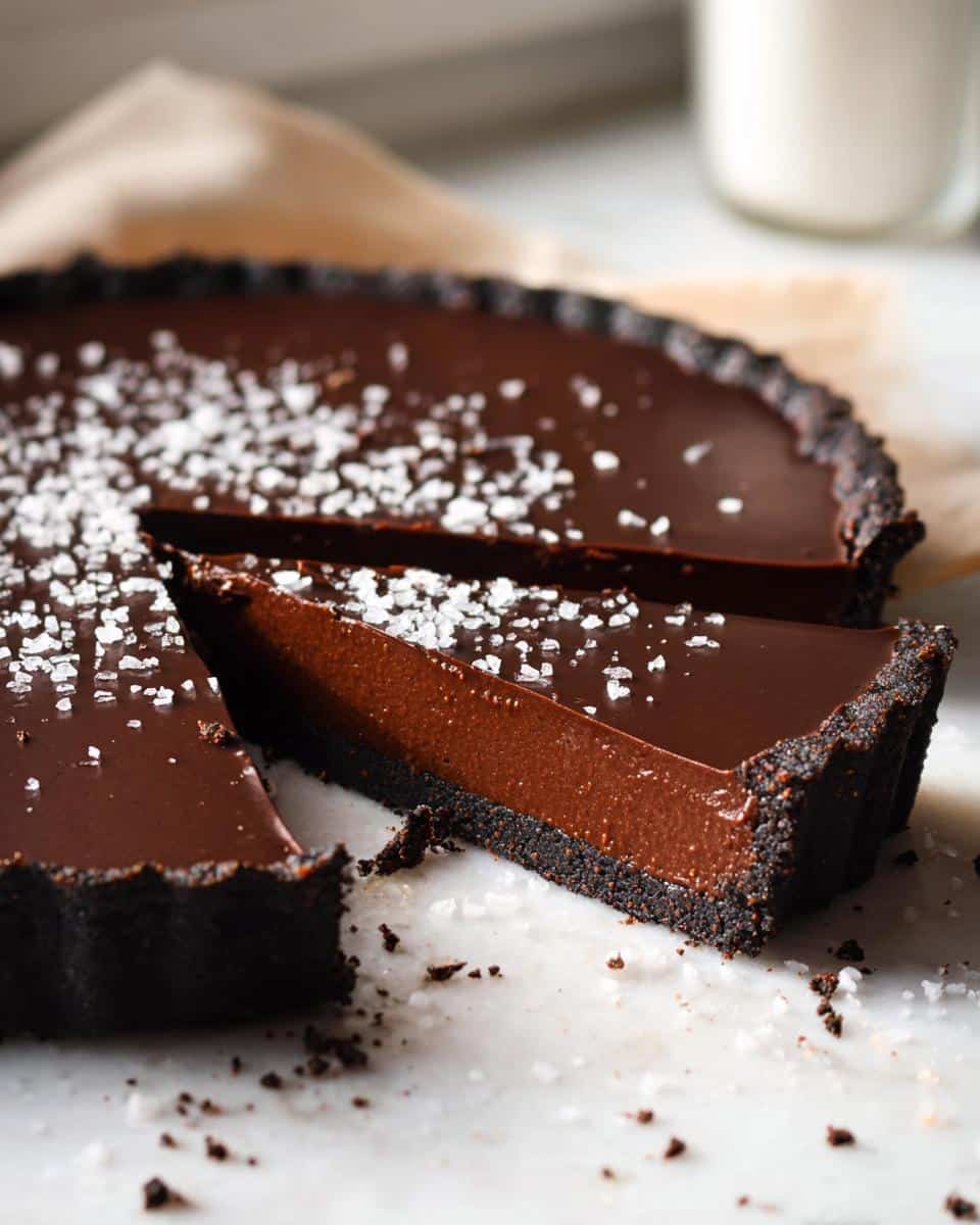 A slice cut from a rich Chocolate Ganache Tart with a dark crust, topped with flaky sea salt.