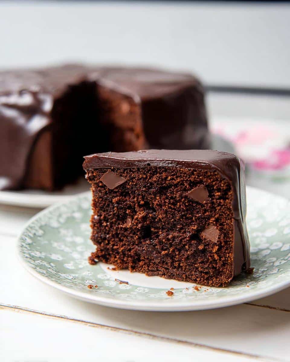 A moist slice of Chocolate Mud Thermomix Cake covered in glossy chocolate ganache, sitting on a patterned plate.