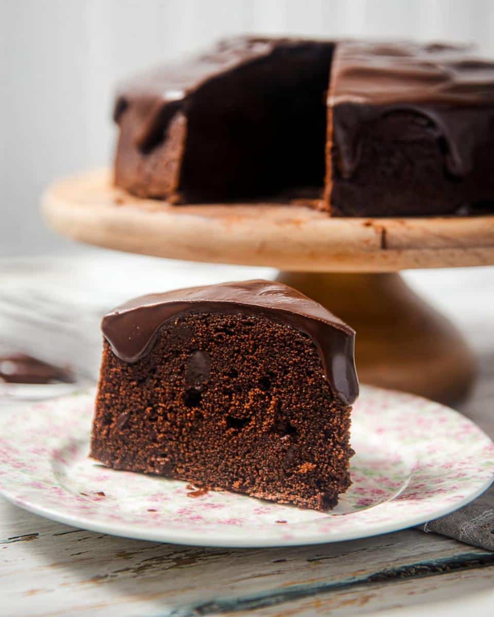 A slice of dense, dark Chocolate Mud Thermomix Cake with glossy chocolate ganache dripping down the side, served on a floral plate.
