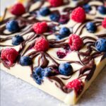 A close-up, overhead view of a piece of frozen Chocolate Yogurt Bark with Berries, featuring white yogurt base, dark chocolate swirls, raspberries, and blueberries.