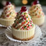 A close-up of one of the Christmas Tree Mini Cheesecakes topped with white frosting, red and green sprinkles, and a cherry.