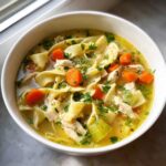 Close-up of a white bowl filled with rich Chicken Noodle Soup, featuring egg noodles, shredded chicken, carrots, and celery.