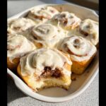 Close-up of freshly baked Classic Gluten-Free Cinnamon Rolls covered in thick white frosting, one roll has a bite taken out.