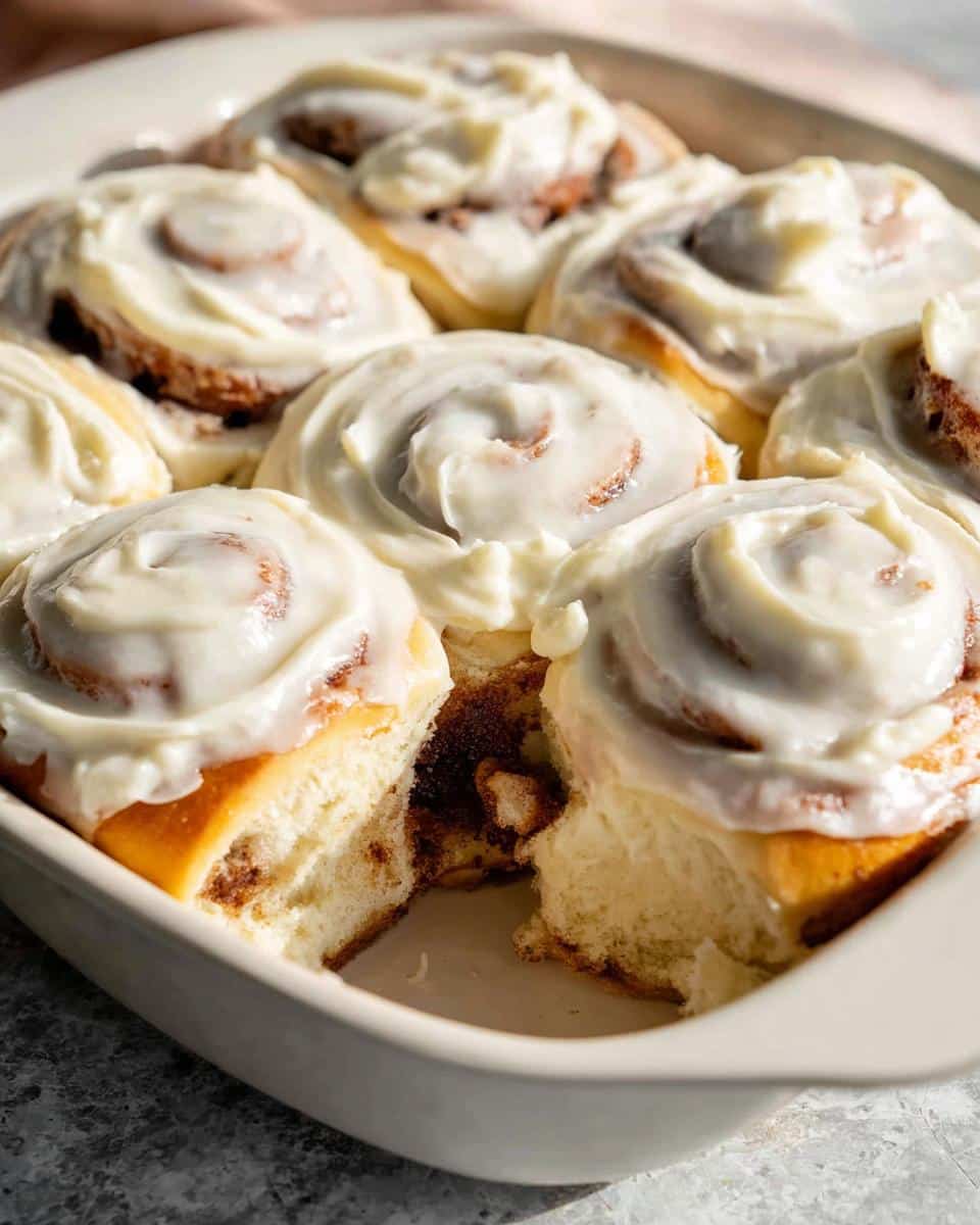 A close-up of freshly baked Classic Gluten-Free Cinnamon Rolls covered in thick white cream cheese frosting in a baking dish.