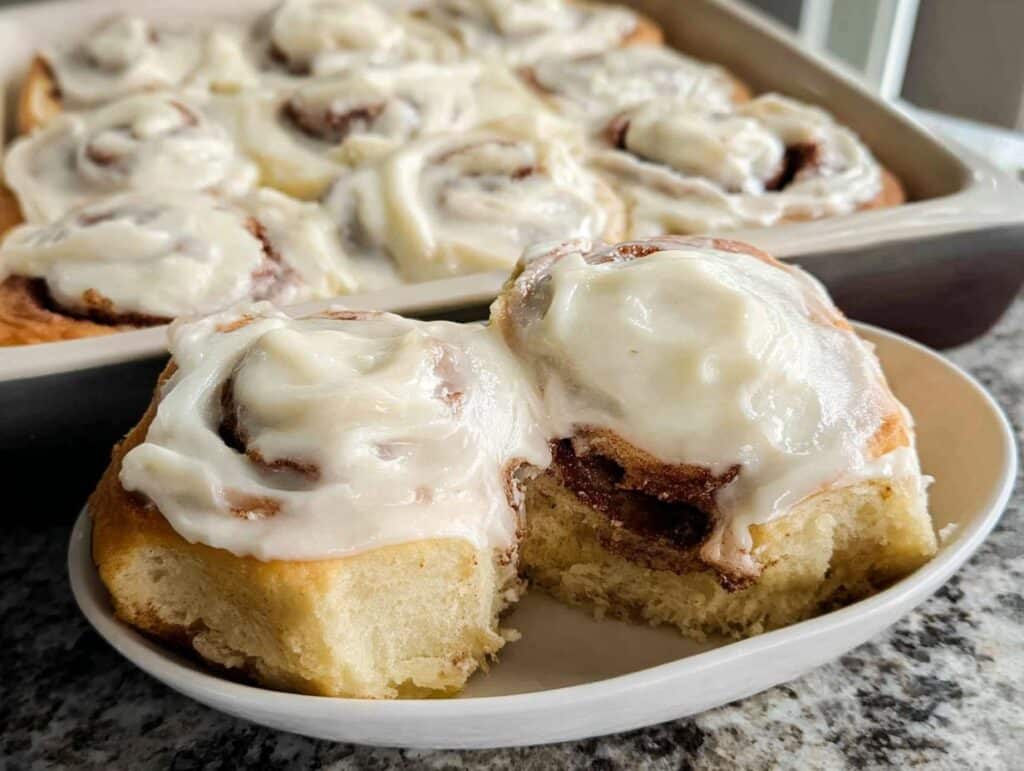 A serving of gooey Classic Gluten-Free Cinnamon Rolls, generously topped with cream cheese frosting, with more rolls visible in the background pan.