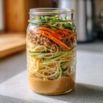 A layered Cold Sesame Peanut Noodle Jar showing sauce on the bottom, noodles, meat, carrots, and green onions on top.