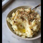 A close-up of a bowl of fluffy Creamy Mashed Potatoes with Butter & Garlic, topped with toasted nuts and herbs.