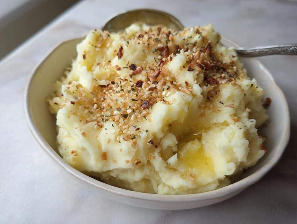 A bowl of fluffy Creamy Mashed Potatoes with Butter & Garlic, topped with toasted garlic bits and herbs.