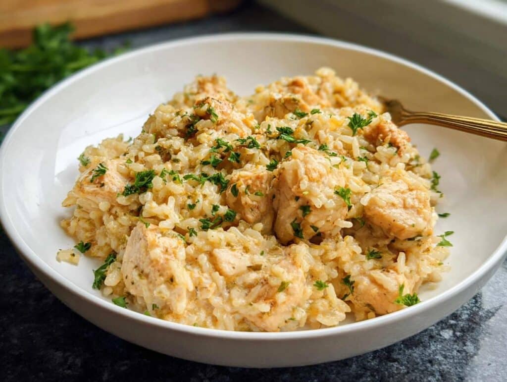 A close-up of a white bowl filled with creamy one-pot chicken and rice, garnished with fresh parsley.