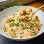 A spoonful of creamy one-pot chicken and rice being lifted from a white bowl, garnished with fresh parsley.