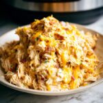 A close-up of shredded chicken coated in a creamy sauce, topped with melted cheddar cheese and bacon bits, representing Creamy Ranch Crack Chicken.