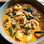 Close-up of a bowl filled with rich, orange Creamy Sausage Tortellini Autumn Soup, featuring tortellini pasta, ground sausage, and spinach.