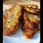 A stack of golden brown, crispy Parmesan crusted chicken cutlets seasoned with herbs on a white plate.
