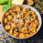 A bowl filled with creamy Dump & Go Chicken Tortellini Crockpot, topped with Parmesan cheese and fresh spinach.