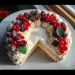 A beautiful easy Christmas Wreath Cake with a slice removed, topped with whipped cream and mixed red and dark berries.
