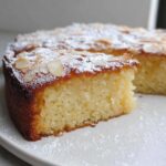 A close-up of a moist slice of Gluten-Free Thermomix Coconut Cake topped with flaked almonds and powdered sugar.
