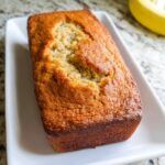 A freshly baked, golden brown loaf of Almond Flour Banana Bread resting on a white rectangular plate.