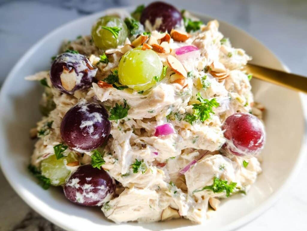 A close-up of creamy Greek Yogurt Chicken Salad topped with red and green grapes, sliced almonds, and fresh parsley.