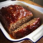 A freshly baked Homestyle Meatloaf with Glaze, sliced in a white ceramic loaf pan.