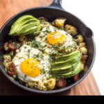 Close-up of a Keto Breakfast Bowl with Cauliflower Hash, topped with two sunny-side-up eggs, sliced avocado, and herbs.