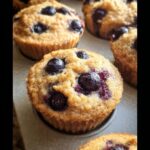 Close-up of freshly baked Keto Coconut Flour Muffins studded with plump blueberries in a muffin tin.