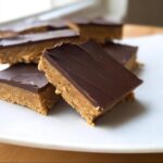Close-up of several rich Keto Peanut Butter Breakfast Bars stacked on a white plate, featuring a thick peanut butter base and a dark chocolate topping.
