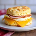 A close-up of a Keto Savory Biscuit Sandwich featuring a fried egg, melted cheese, and ham between two fluffy biscuits.