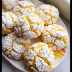 A close-up of several bright yellow Lemon Crinkle Cookies heavily coated in cracked white powdered sugar, arranged on a white plate.