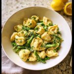 A white bowl filled with creamy Lemon Garlic Tortellini with Peas and fresh herbs, with lemons visible in the background.