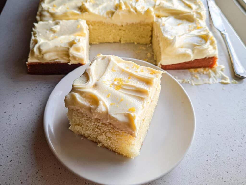 A square slice of moist Lemon Yogurt Sheet Cake topped with thick, swirled white frosting and lemon zest.