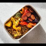 Overhead view of a stainless steel lunch box filled with lentils, roasted broccoli, peppers, and sliced sausage, perfect for a Lentil & Roasted Vegetable Lunch Box.