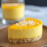 A slice of bright yellow Mango Mousse Thermomix Cups dessert with a biscuit base and coconut flakes.