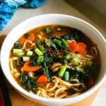 A close-up of a white bowl filled with Miso Vegetable Soup featuring noodles, carrots, dark greens, and scallions.